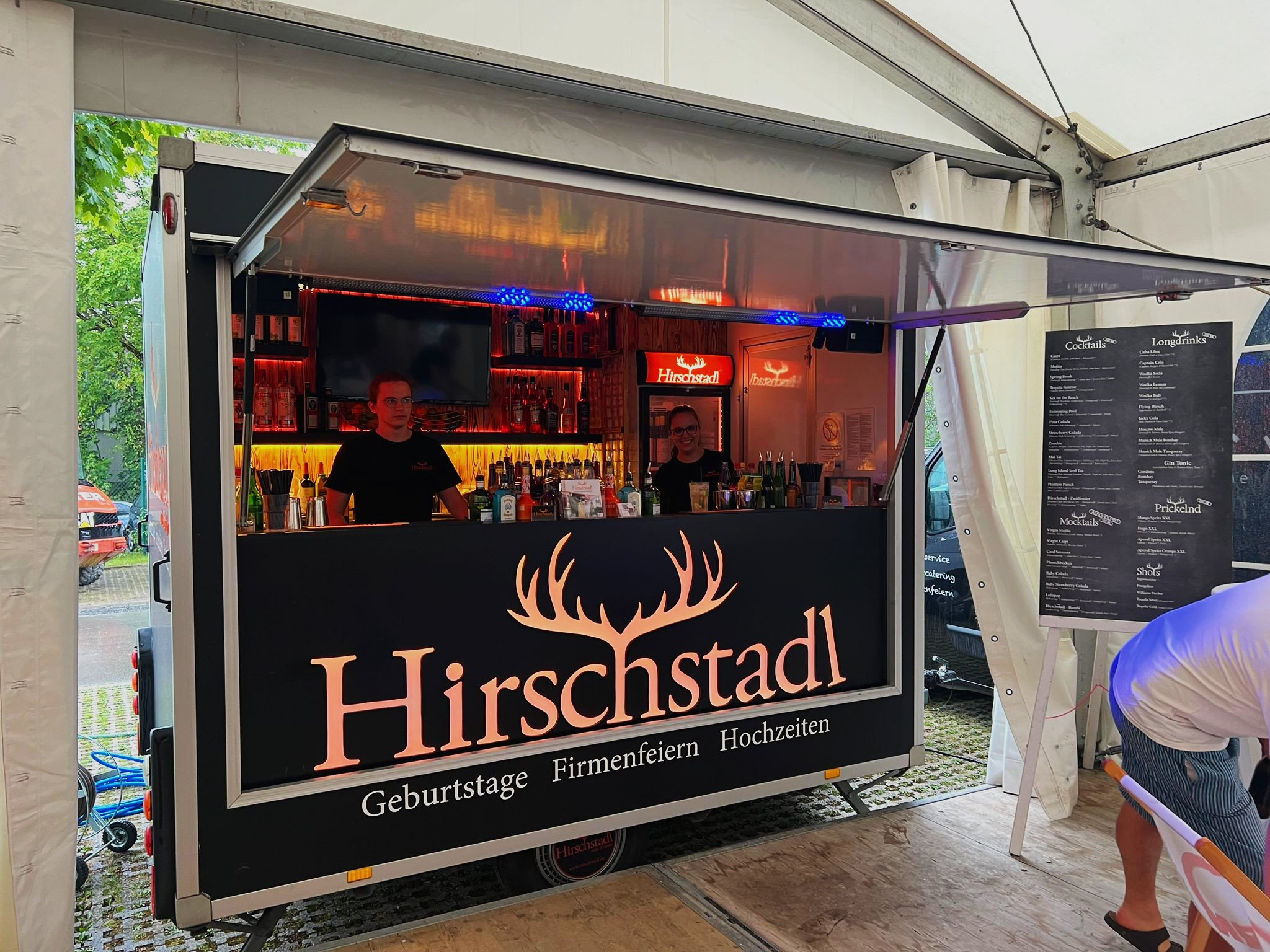 Geoeffneter BarWagen im Zelt, zwei Barkeeper an der Ausgabe, darueber das beleuchtete Hirschstadl-Schild